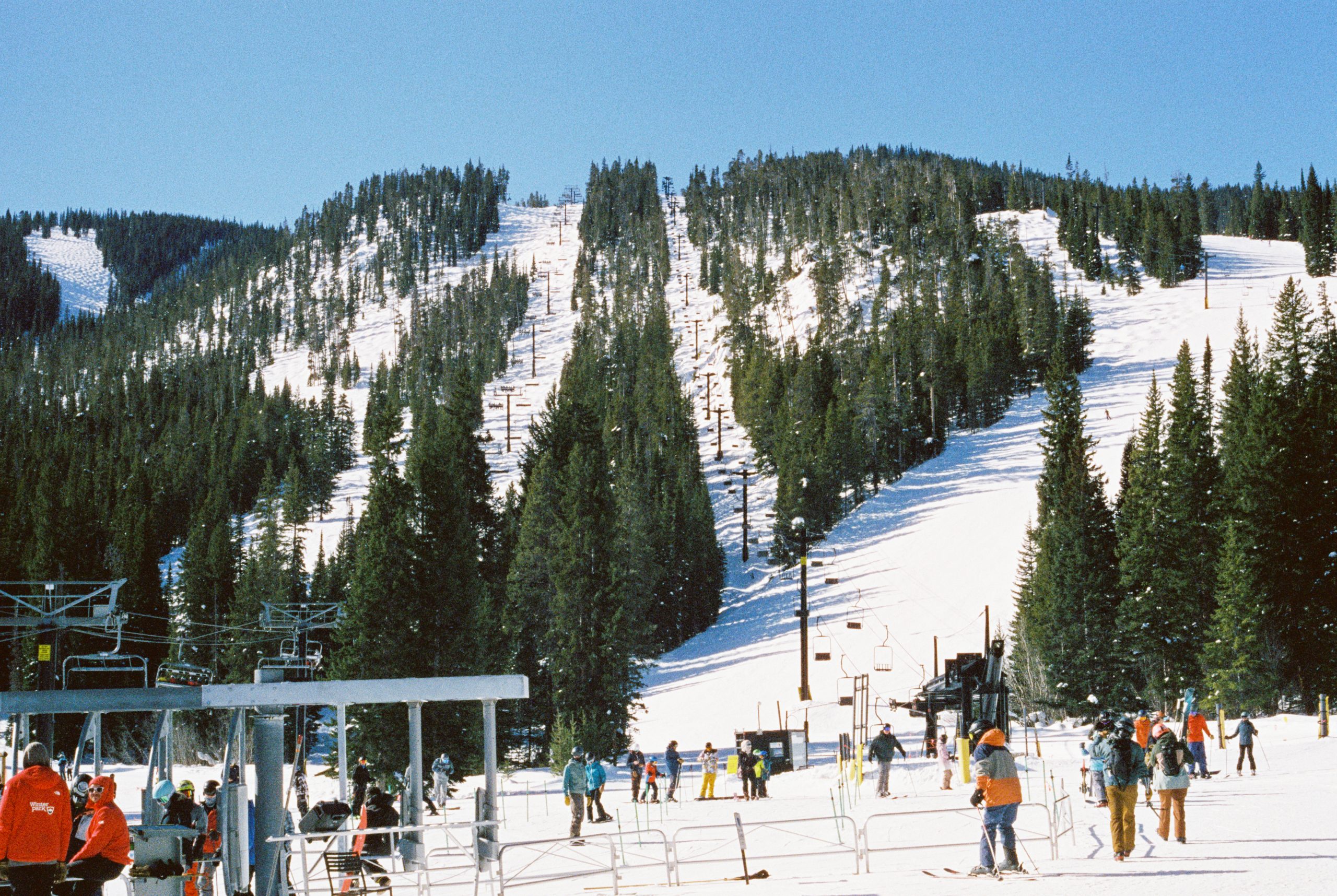 Skiers gather at the base of a snowy mountain, surrounded by pine trees and ski lifts under a clear blue sky. The scene captures the charm of film photography, with people preparing to ski while others wait near the lift area. | Photo by Millworks Film Photography & Prints