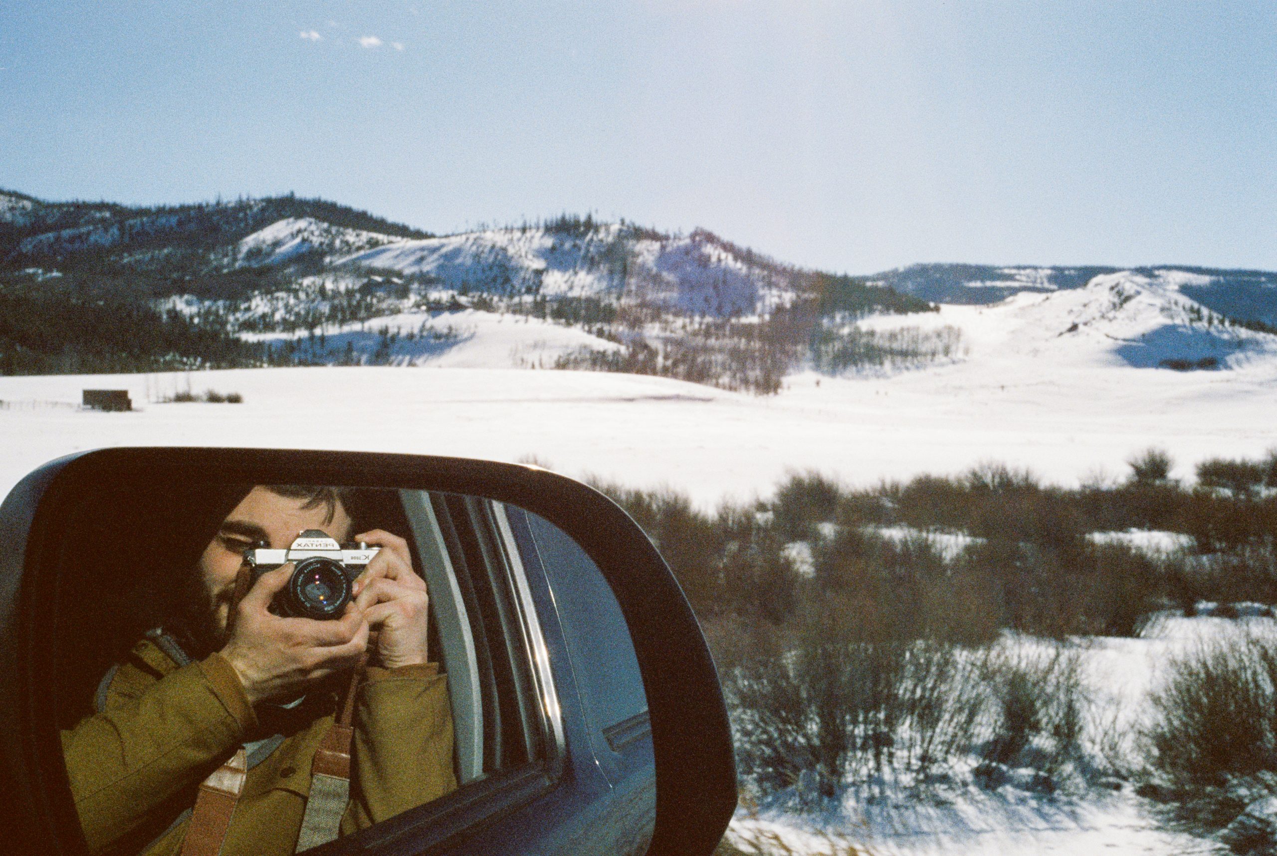A person in a car takes a film photo with a camera, reflected in the side mirror. Snow-covered hills and sparse bushes appear outside under a clear blue sky, capturing the essence of professional photography. | Photo by Millworks Film Photography & Prints