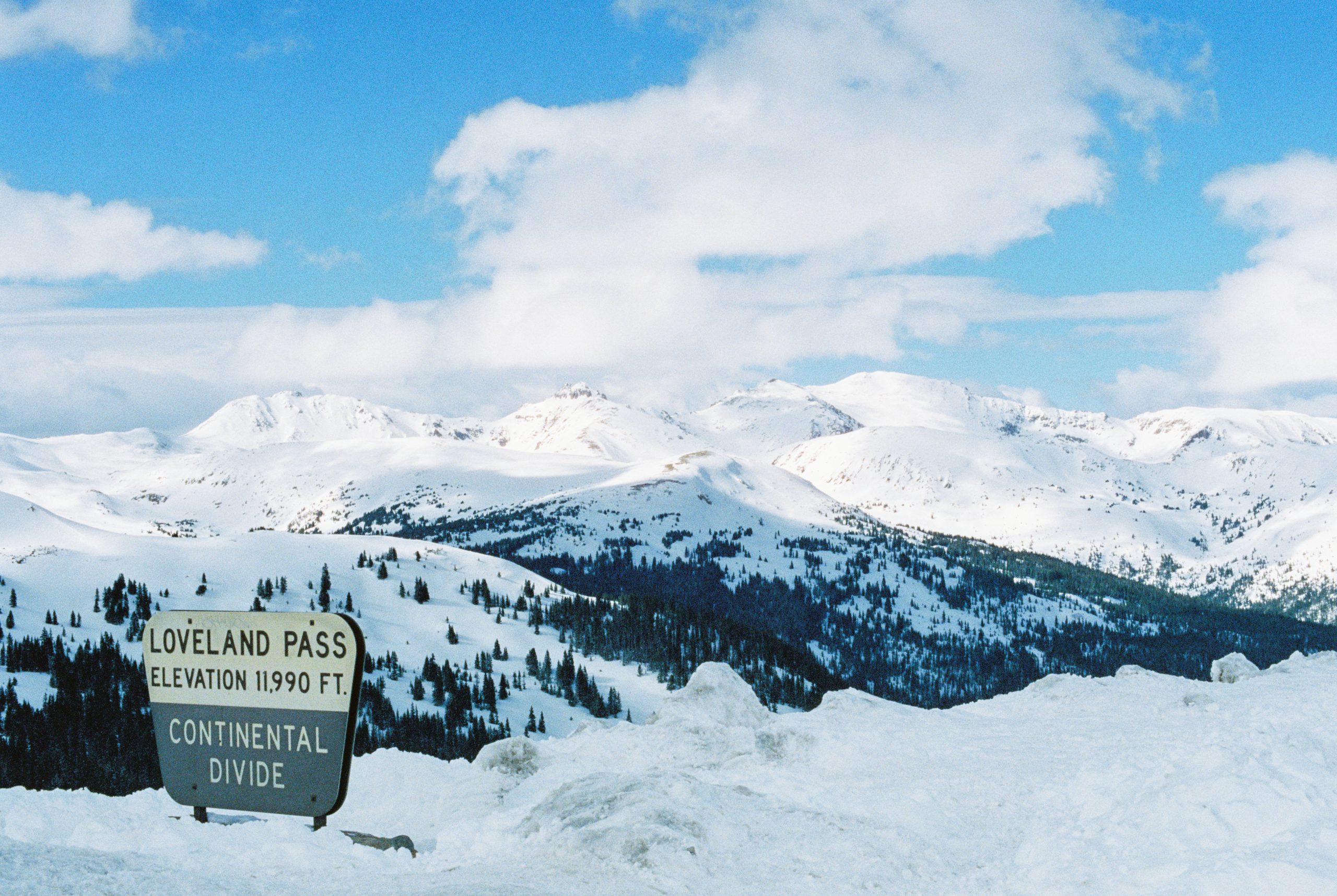 A sign reads Loveland Pass Elevation 11,990 ft. Continental Divide in front of snow-covered mountains and blue sky with clouds. Pine trees dot the slopes, captured in crisp professional photography. | Photo by Millworks Film Photography & Prints
