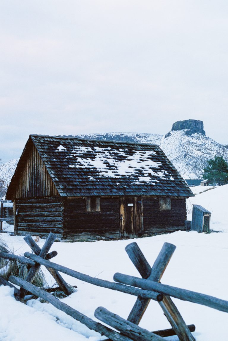 A rustic wooden cabin with a snow-covered roof sits in a snowy landscape, surrounded by a wooden fence. Captured as a stunning film photo, a butte rises against the cloudy sky in the background. | Photo by Millworks Film Photography & Prints