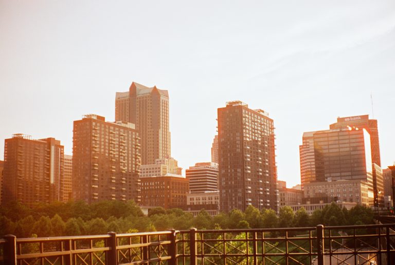 A city skyline with mid- and high-rise buildings under a bright sky, sunlight casting a warm glow. Trees and a decorative metal fence are in the foreground, captured with the nostalgic charm of film photography. | Photo by Millworks Film Photography & Prints