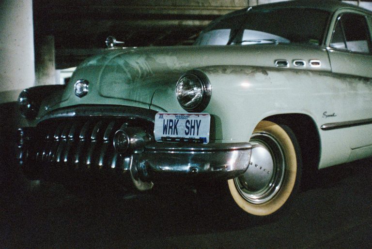 A vintage pale green car with chrome bumper and round headlight is parked indoors. Captured in a film photo, the license plate reads WRK SHY. The car has whitewall tires and a Special badge on the side. | Photo by Millworks Film Photography & Prints