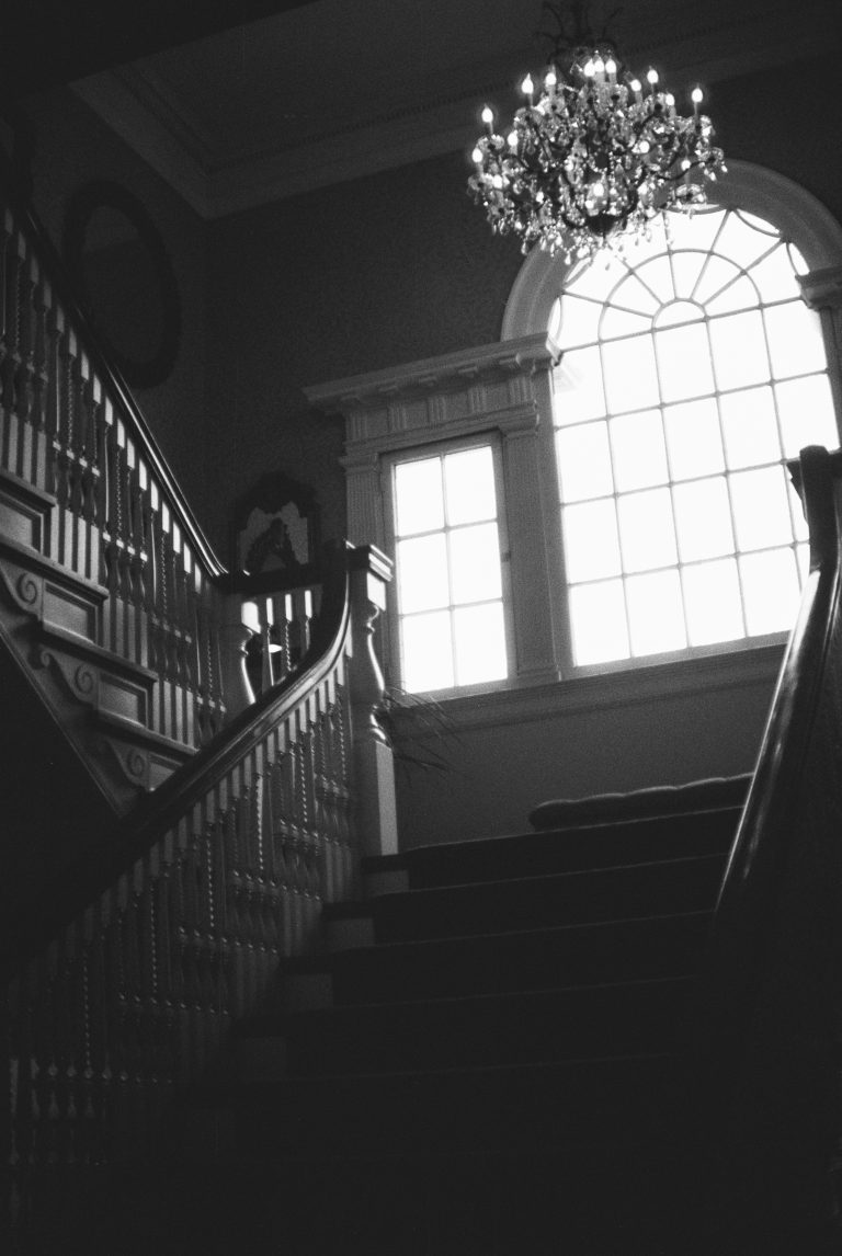 A grand staircase in a dimly lit interior, with ornate railings, a large arched window casting bright light, and a chandelier overhead—all beautifully captured in the style of film photography. | Photo by Millworks Film Photography & Prints
