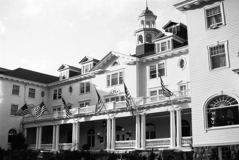 A large, historic white building with a columned porch, multiple American flags hanging, a central cupola, and many windows; captured in classic black and white film photography. | Photo by Millworks Film Photography & Prints