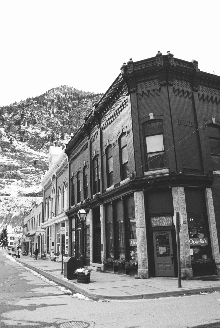 A black-and-white film photo of a historic brick building on a quiet street, with snow-dusted mountains in the background. The building has large windows and ornate architectural details, capturing the charm of film photography. | Photo by Millworks Film Photography & Prints