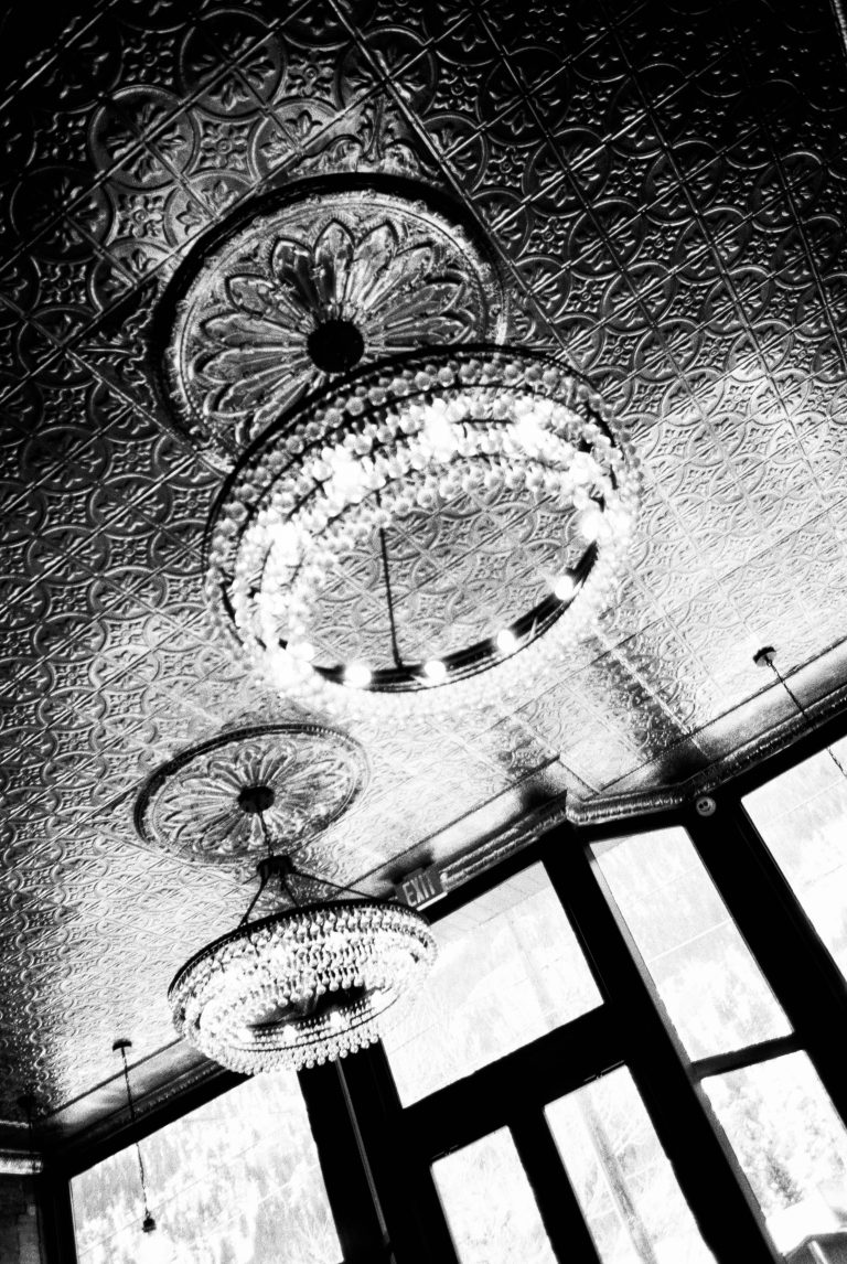 Black and white film photography captures ornate chandeliers hanging from an intricately patterned metallic ceiling, with large windows and natural light illuminating the background. | Photo by Millworks Film Photography & Prints
