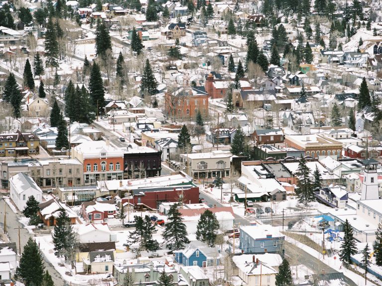 A small town seen from above, covered in snow. Streets, houses, and buildings are surrounded by evergreen trees, creating a wintry scene—like a film photo with rooftops blanketed in white. | Photo by Millworks Film Photography & Prints