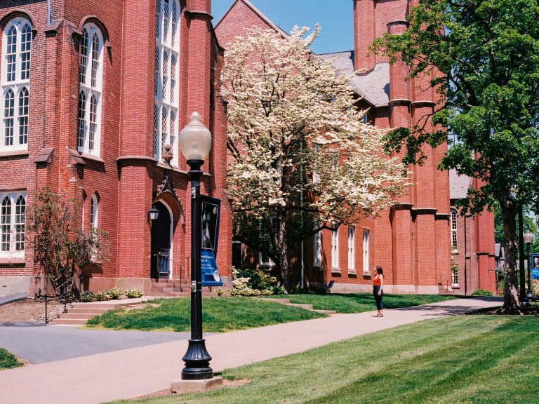 A person in red stands on a sidewalk near a flowering tree outside a large red-brick building with tall windows on a sunny day. Captured as a film photo, the image features a streetlamp and blue banner in the foreground. | Photo by Millworks Film Photography & Prints