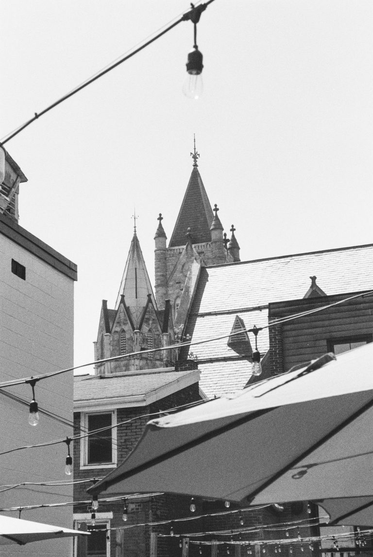 Black and white film photography captures a church steeple and pointed rooftops behind buildings and large patio umbrellas, with string lights hanging overhead. | Photo by Millworks Film Photography & Prints