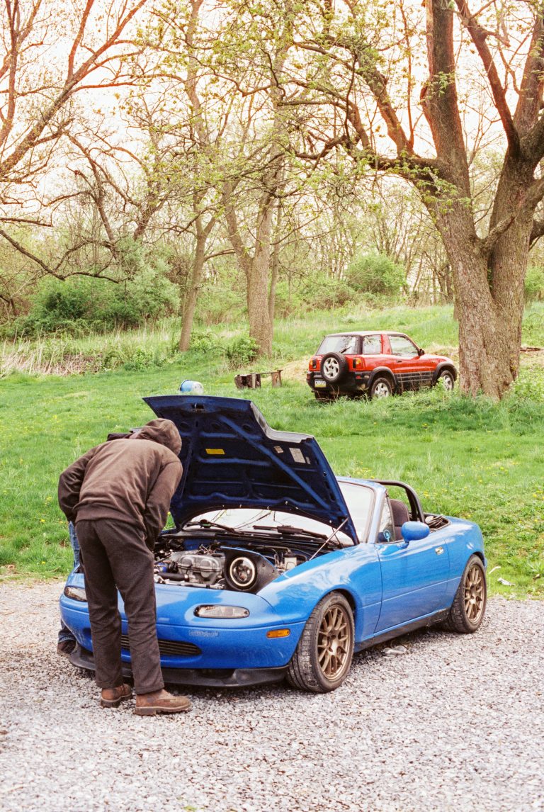 A person in dark clothing inspects the engine of a blue convertible with the hood open, captured in a film photo on gravel near grass and trees, with a red SUV visible in the background. | Photo by Millworks Film Photography & Prints