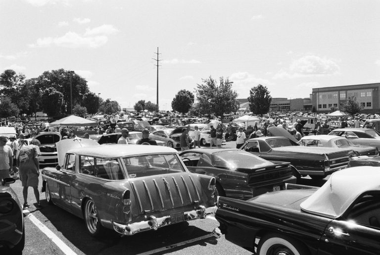 A busy outdoor car show in a parking lot showcases many classic cars and people strolling on a sunny day, with trees, tents, and a building in the background. Captured as a striking black and white film photo. | Photo by Millworks Film Photography & Prints