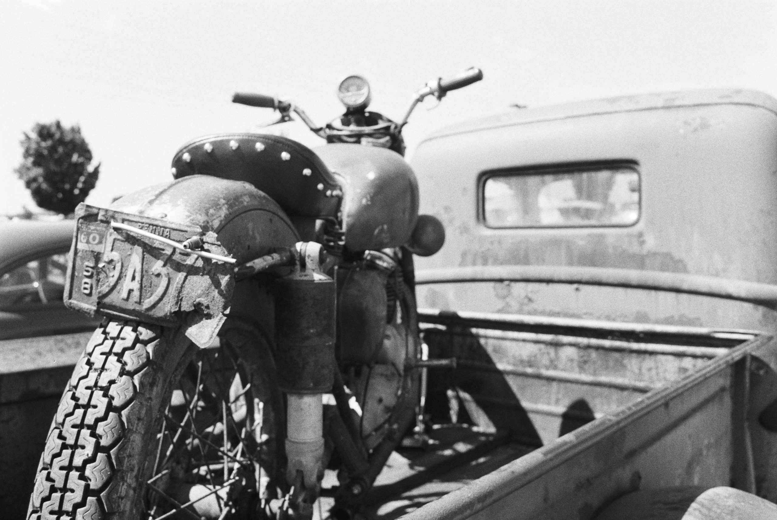 A vintage motorcycle with a worn license plate sits in the bed of an old pickup truck. Captured as a black and white film photo, the image exudes nostalgia and classic style, with another vehicle and a distant tree in the background. | Photo by Millworks Film Photography & Prints