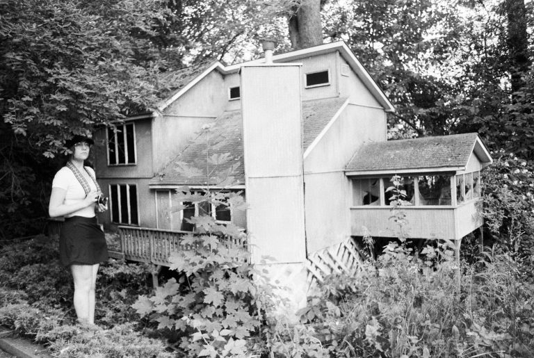 A person holding a camera stands next to a large, detailed model of a house surrounded by overgrown plants and trees in a wooded area. Captured in black and white, this film photography image evokes a timeless, nostalgic atmosphere. | Photo by Millworks Film Photography & Prints
