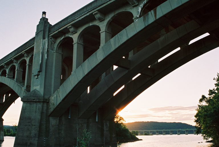 A concrete arch bridge stretches over a river at sunset, the sun peeking through its arches. Captured in the warm tones of film photography, trees and hills fade into the backdrop while another distant bridge completes the scene. | Photo by Millworks Film Photography & Prints