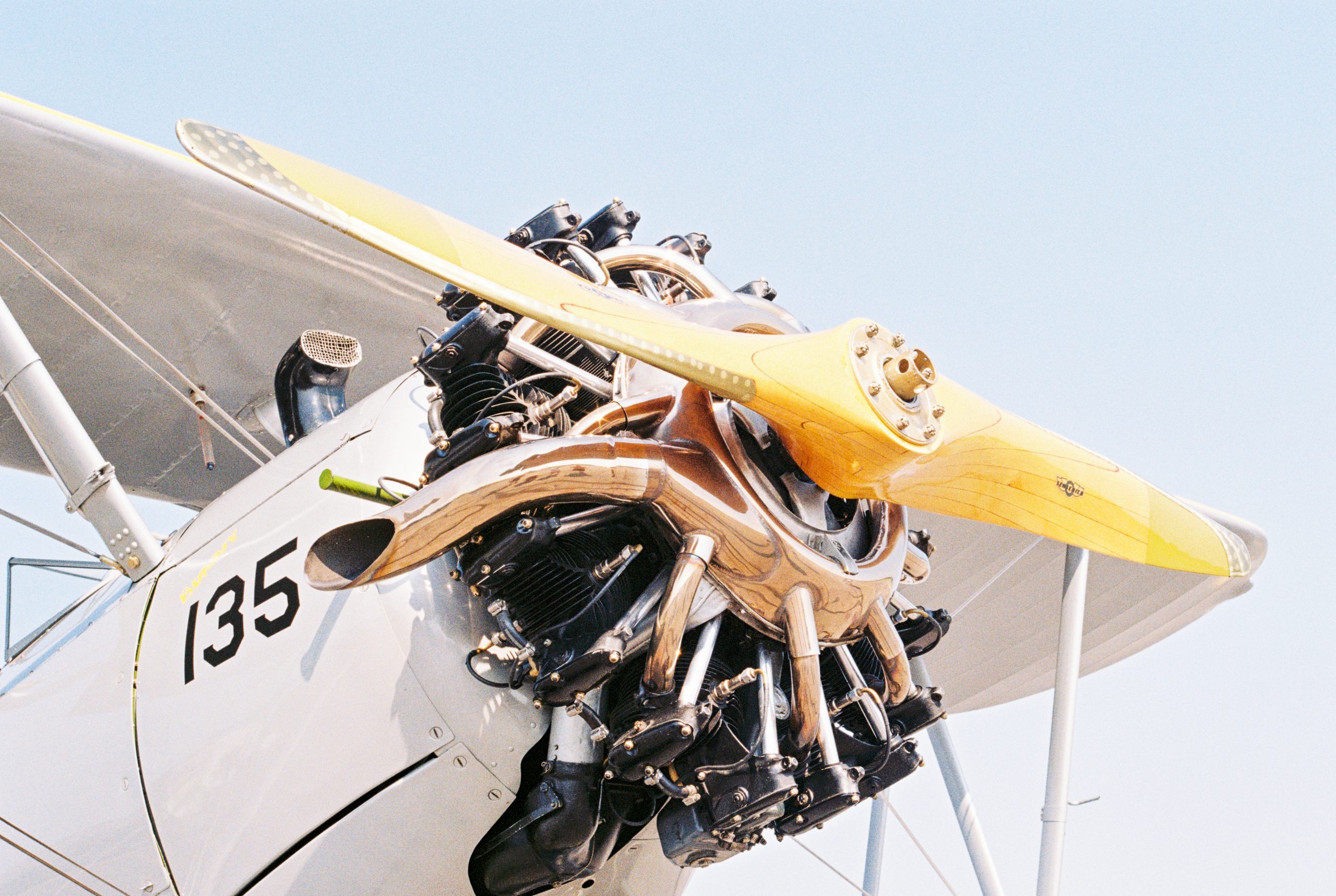 Close-up of a vintage biplane’s yellow wooden propeller and exposed radial engine, captured in rich detail like a classic film photo. Part of the white wing and fuselage is visible against a clear blue sky, with the number 135 painted on the aircraft. | Photo by Millworks Film Photography & Prints