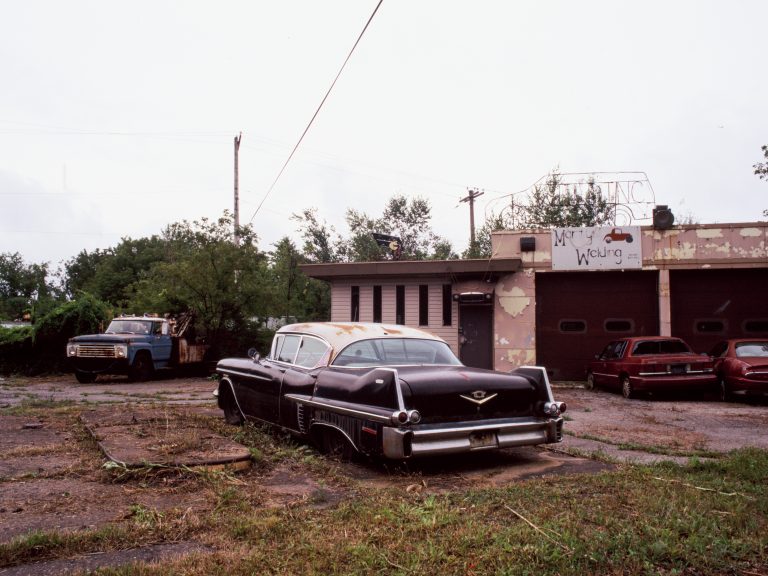 An old black classic car is parked on a cracked, overgrown lot near a weathered auto repair shop with faded paint. Captured as a film photo, the scene includes a blue pickup and two red cars, all framed by trees and cloudy skies. | Photo by Millworks Film Photography & Prints