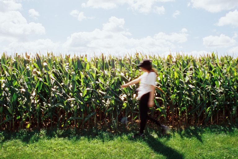 A person in a hat and white shirt walks in front of a tall, green cornfield under a bright, partly cloudy sky. Captured as a film photo, the blurred figure suggests movement and adds an artistic touch to this serene rural scene. | Photo by Millworks Film Photography & Prints