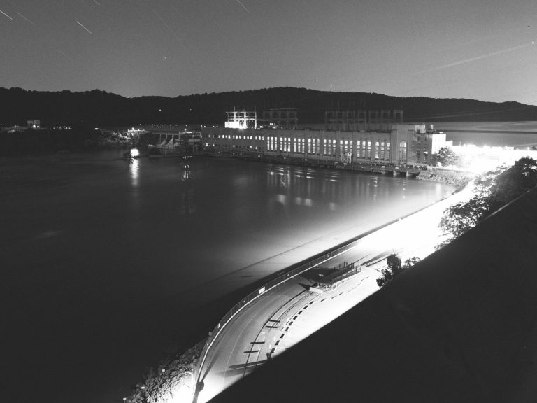 Black and white night view of a hydroelectric dam with a large building, calm water, and curved road in the foreground; hills and faint star trails in the background evoke classic film photography and professional photography techniques. | Photo by Millworks Film Photography & Prints