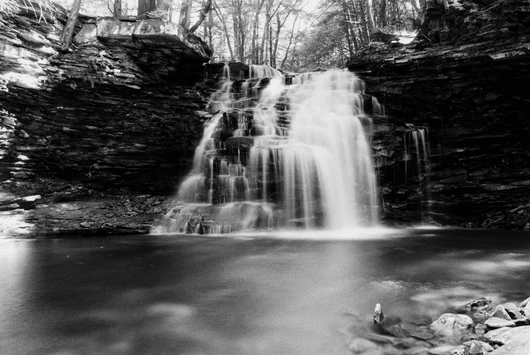 A black and white film photo of a waterfall cascading over rocky ledges into a calm pool, surrounded by trees and rugged cliffs. The water appears smooth, creating a soft, misty effect. | Photo by Millworks Film Photography & Prints