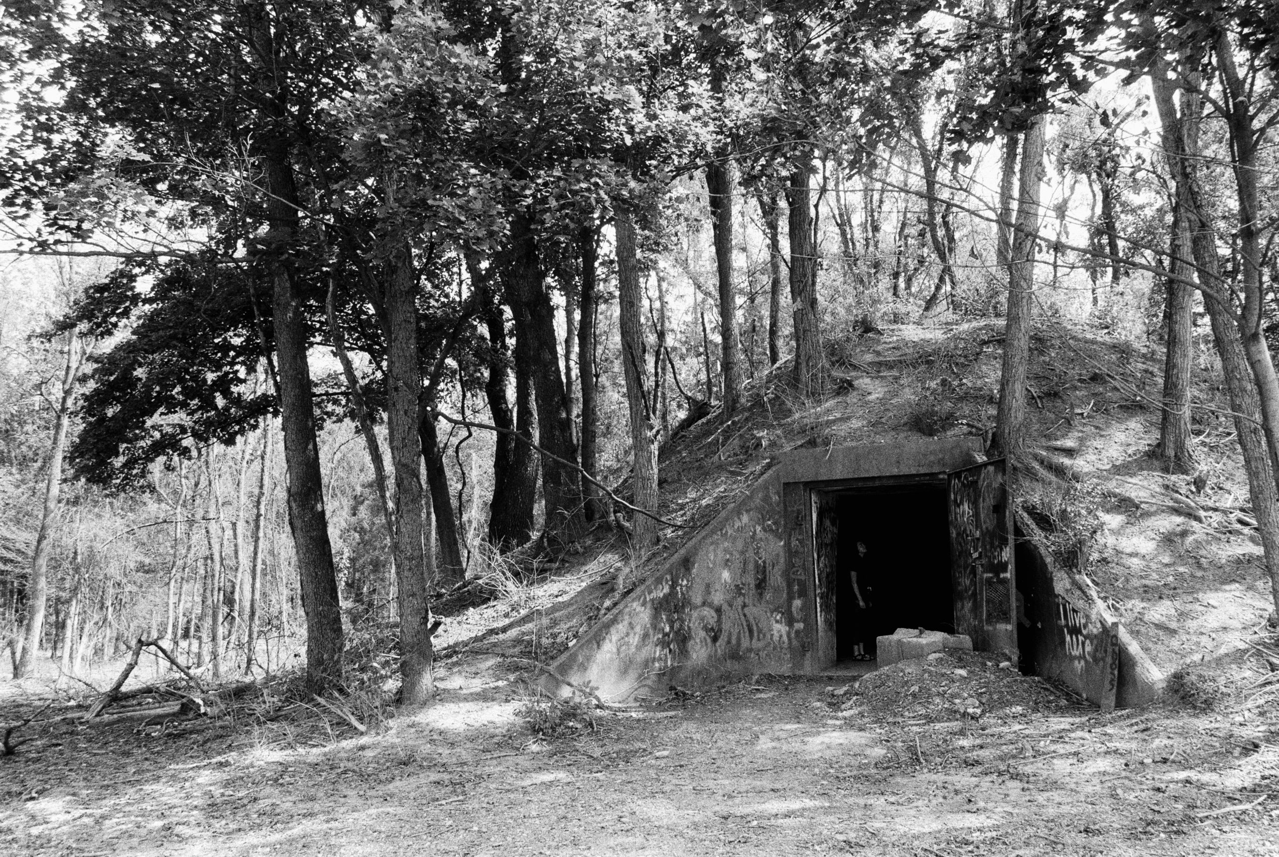 A black and white film photo of a forest with an old, concrete bunker built into a small hill. The entrance is dark and partially overgrown, with trees and shadows enhancing the moody feel of film photography. | Photo by Millworks Film Photography & Prints