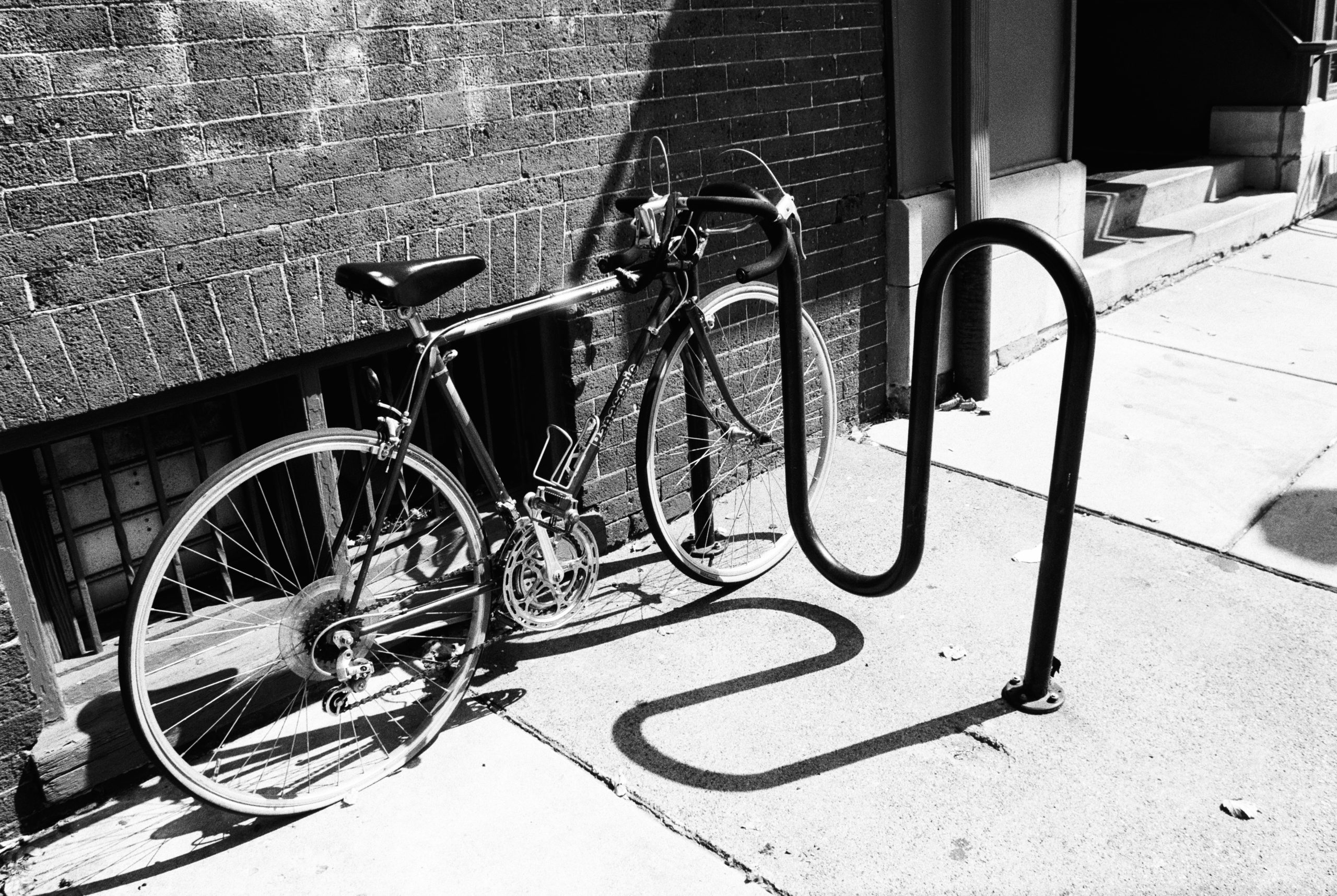 A bicycle is parked and locked to a curved metal bike rack on a sidewalk next to a brick building. Sunlight casts strong shadows on the pavement, evoking the timeless charm of film photography. | Photo by Millworks Film Photography & Prints