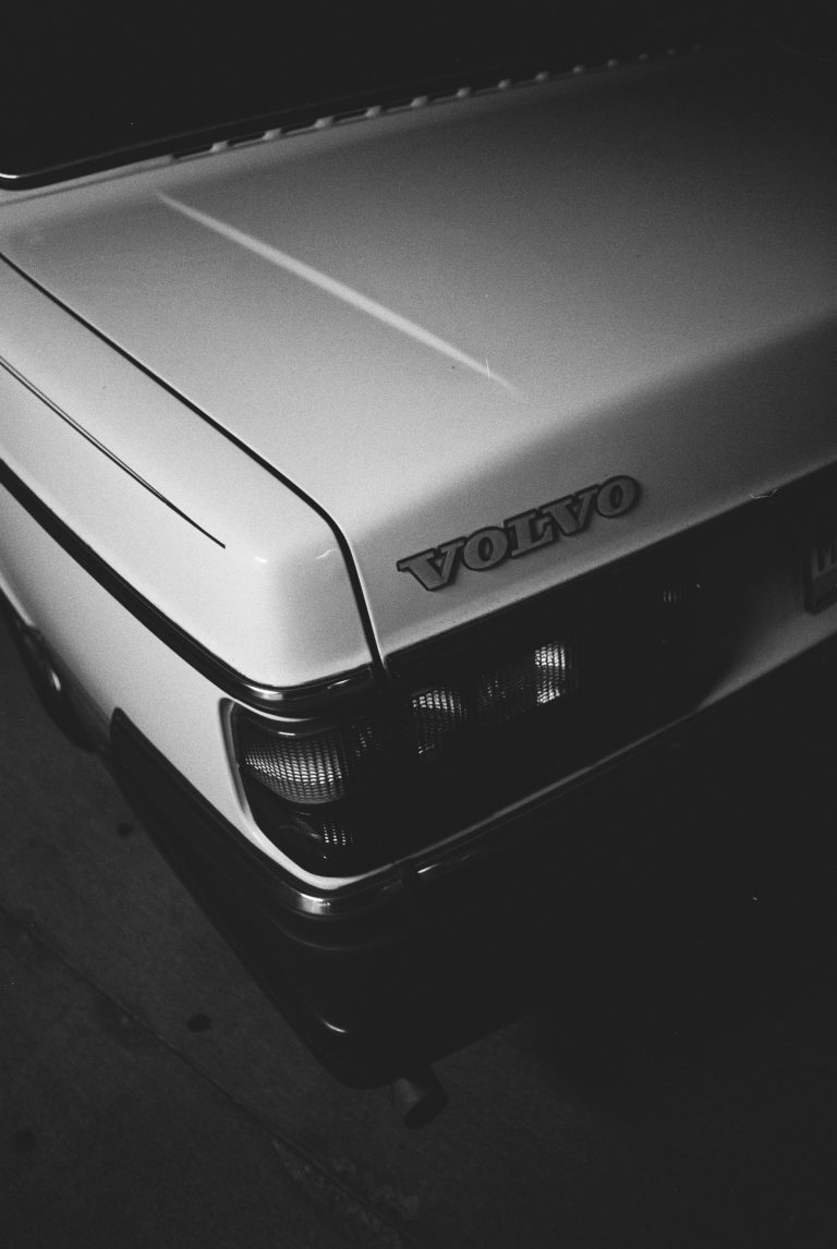 Black and white film photo showing the rear corner of a white Volvo car, with the VOLVO badge visible above the tail lights. Captured at an angle, this film photography image highlights the trunk and part of the bumper. | Photo by Millworks Film Photography & Prints
