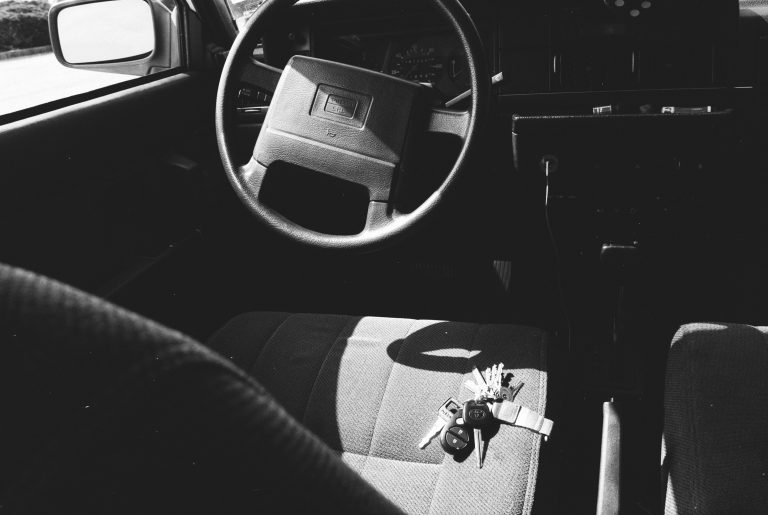 Black and white film photo of a car’s interior, showing the steering wheel and front seat. A set of car keys and a watch rest on the fabric seat in sunlight, capturing the timeless touch of film photography. | Photo by Millworks Film Photography & Prints