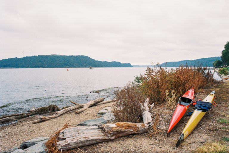 Two kayaks, one red and one yellow, rest on a rocky, driftwood-strewn shoreline beside calm water. Captured in the rich tones of film photography, distant hills and a boat complete this peaceful scene under a cloudy sky. | Photo by Millworks Film Photography & Prints