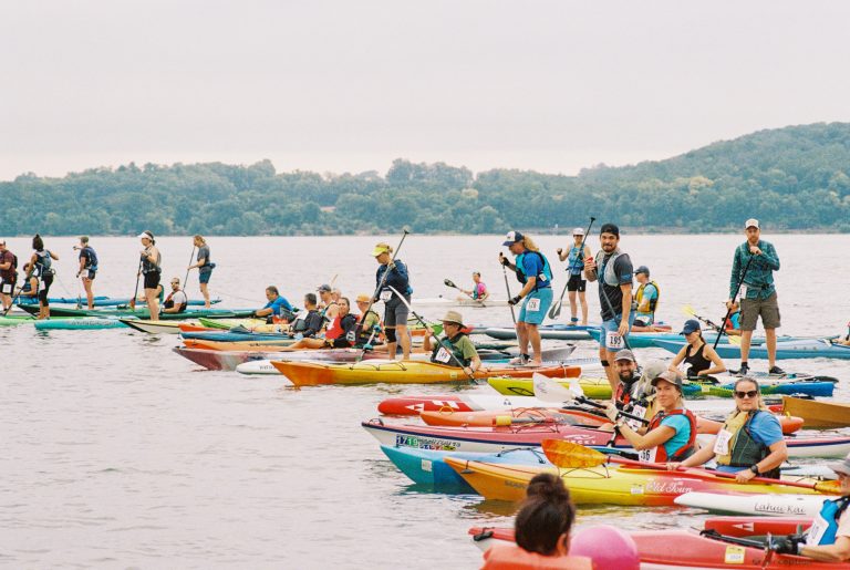 A group of people in colorful kayaks and paddleboards gather on a calm lake, wearing life vests and holding paddles, with wooded hills in the background—captured in vivid detail through professional photography. | Photo by Millworks Film Photography & Prints