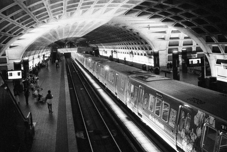 A black-and-white film photo of a subway station with an arched, geometric ceiling captures people waiting or walking near a stopped train. Illuminated signs glow above, giving this grainy scene a timeless, professional photography feel. | Photo by Millworks Film Photography & Prints