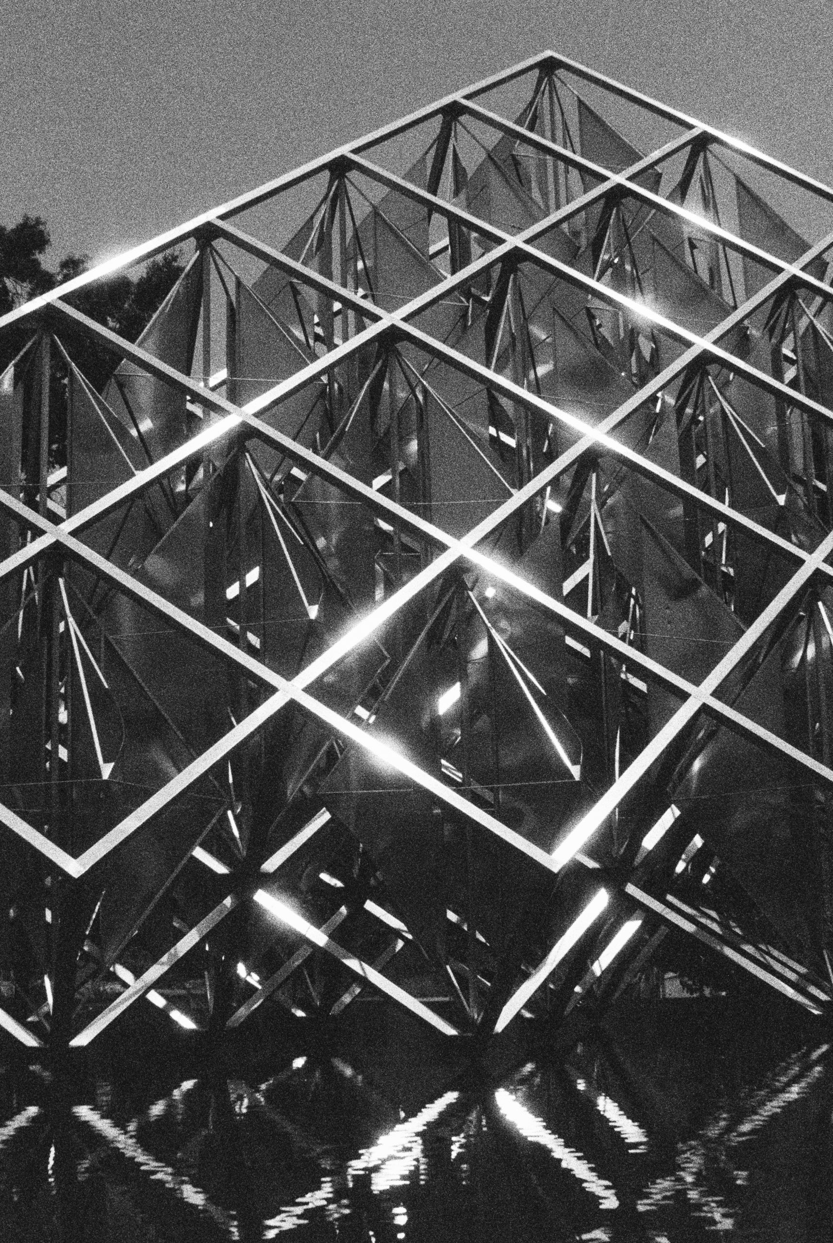A black and white film photo of a geometric glass and metal structure with a lattice design, reflecting light and water below. The structure features intersecting diagonal beams and striking angular shapes. | Photo by Millworks Film Photography & Prints