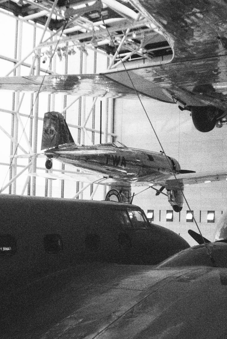Black and white film photo of several vintage airplanes displayed indoors at a museum, with one silver plane suspended above the others, marked with TWA on its side. | Photo by Millworks Film Photography & Prints