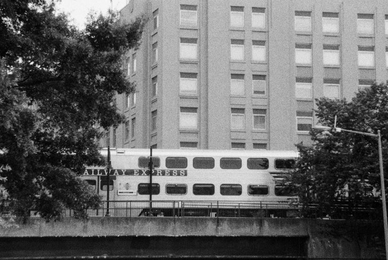 A black and white film photo shows a train labeled “RAILWAY EXPRESS” passing in front of a tall building with many windows, partially obscured by trees. | Photo by Millworks Film Photography & Prints