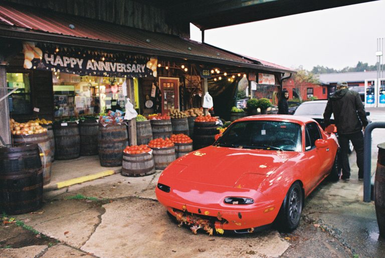 A red sports car is parked in front of a market decorated with barrels of produce and a Happy Anniversary banner. Captured on film photography, two people stand near the car under the awning on a cloudy day. | Photo by Millworks Film Photography & Prints
