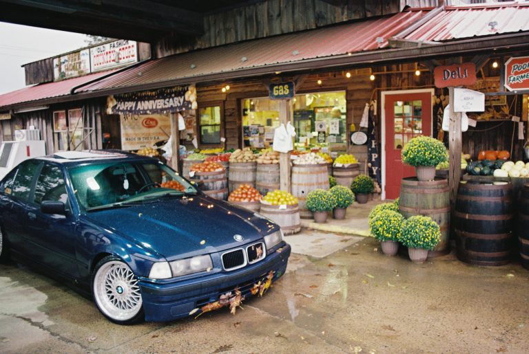 A dark blue BMW with a garland on its bumper sits before a rustic market store, captured in rich film photography. Flower pots, barrels, and vibrant produce line the scene, while a person stands by the deli sign inside. | Photo by Millworks Film Photography & Prints