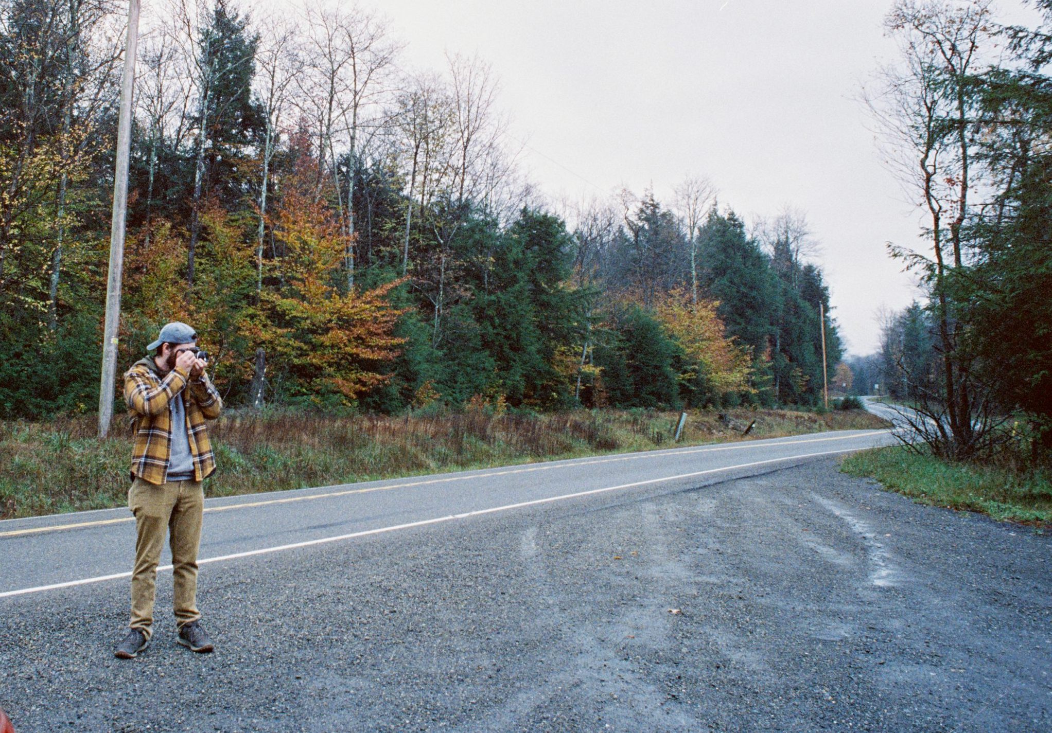 A person wearing a plaid shirt and hat stands beside a quiet paved road, taking a film photo with a camera. The background features trees with autumn foliage and a cloudy sky. | Photo by Millworks Film Photography & Prints
