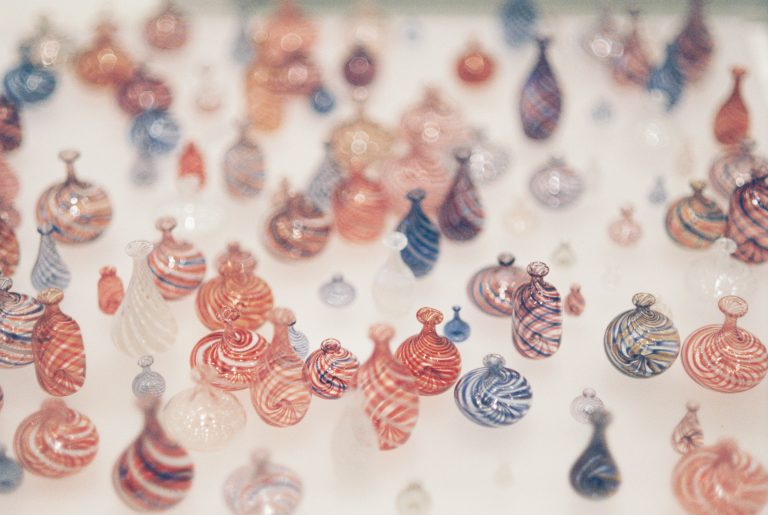 A collection of small, colorful glass vases with swirling patterns in red, white, and blue, arranged closely together on a light surface. The dreamy focus gives the image a nostalgic film photography feel. | Photo by Millworks Film Photography & Prints