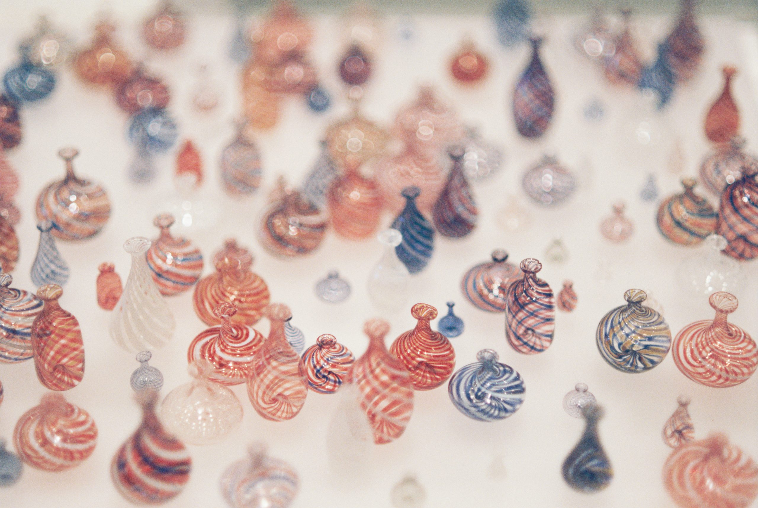 A collection of small, colorful glass vases with swirling patterns in red, white, and blue, arranged closely together on a light surface. The dreamy focus gives the image a nostalgic film photography feel. | Photo by Millworks Film Photography & Prints