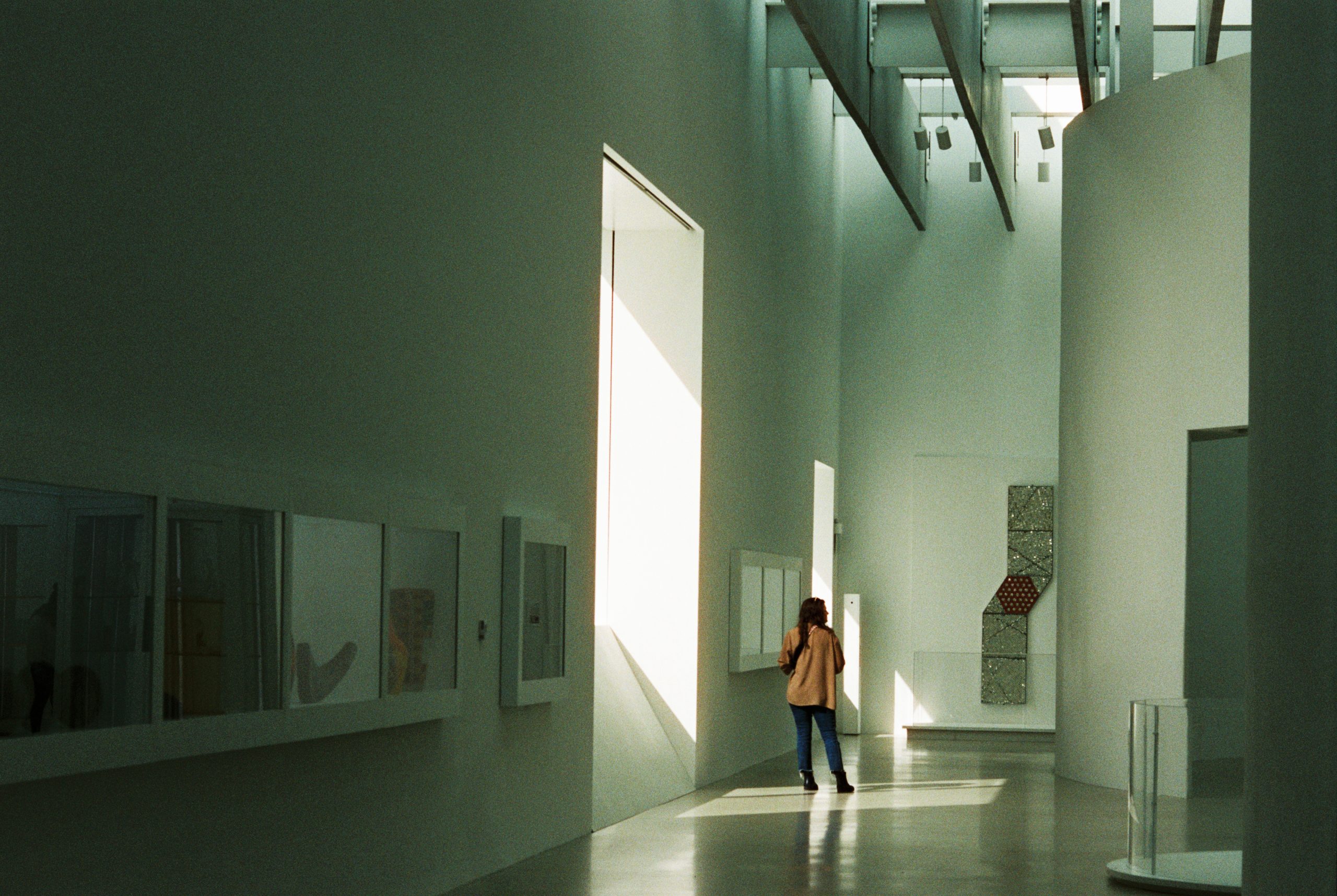 A person stands alone in a modern, minimalist art gallery lit by natural sunlight, reminiscent of a film photography aesthetic that highlights both the white walls and the displayed artwork. | Photo by Millworks Film Photography & Prints