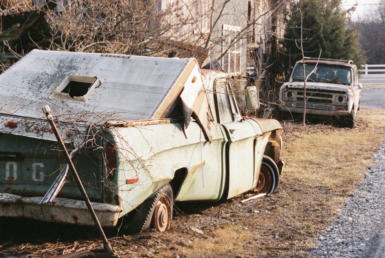 An old, rusted pickup truck with a broken camper top sits abandoned on grass beside a gravel road; captured in this atmospheric film photography style, another rusty truck is visible in the background near some trees. | Photo by Millworks Film Photography & Prints