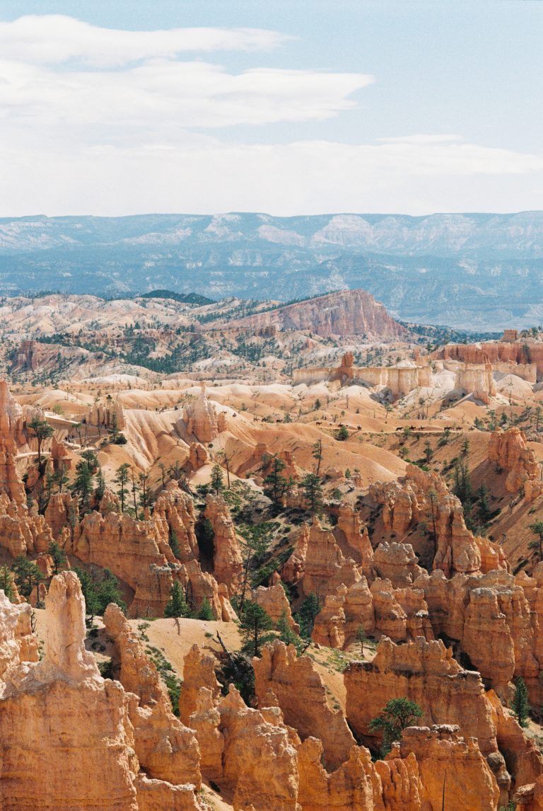 A panoramic view of Bryce Canyon with orange rock spires, scattered trees, and distant mountain ranges beneath a partly cloudy sky, captured in stunning detail through professional photography. | Photo by Millworks Film Photography & Prints