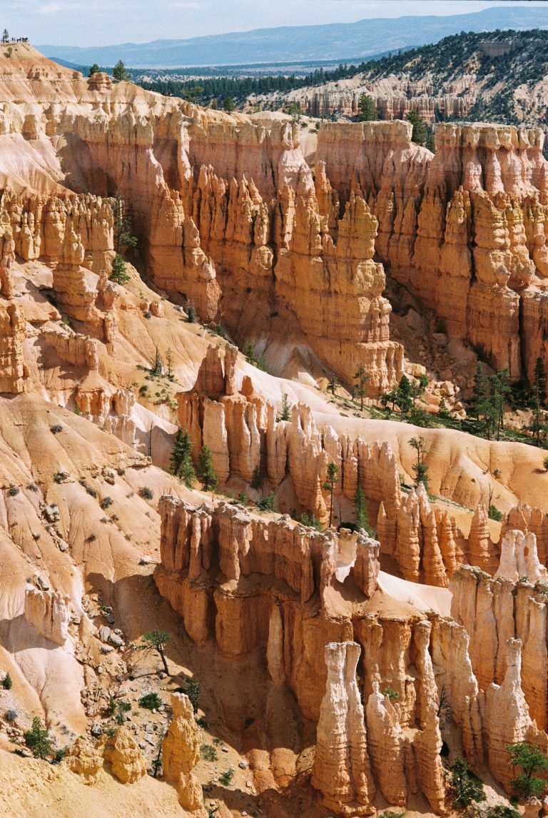 Orange and pink rock formations, known as hoodoos, rise dramatically from the canyon floor at Bryce Canyon National Park under a partly cloudy sky, with scattered green pines dotting the landscape in this striking film photography capture. | Photo by Millworks Film Photography & Prints