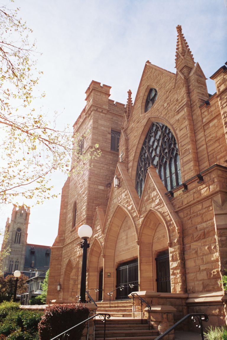 A large stone church with pointed arches, stained glass windows, and gothic architectural details, captured in stunning film photography against a bright sky with trees and another church visible in the background. | Photo by Millworks Film Photography & Prints