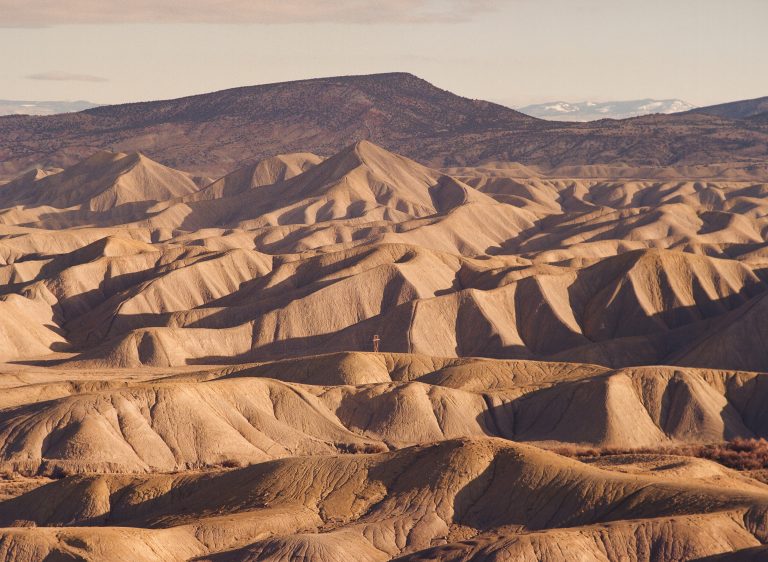 A vast landscape of sunlit, rolling, barren hills with ridged surfaces stretches into the distance, captured in a film photo, with a darker mountain and snow-capped peaks visible in the background under a hazy sky. | Photo by Millworks Film Photography & Prints
