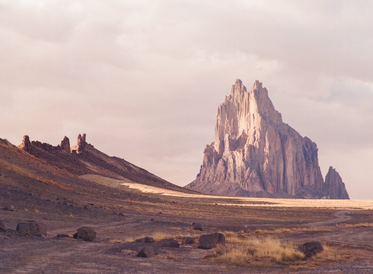 A tall, jagged rock formation rises from a flat, arid landscape under a hazy sky, captured in striking detail with professional photography; scattered boulders and sparse dry grass fill the foreground. | Photo by Millworks Film Photography & Prints
