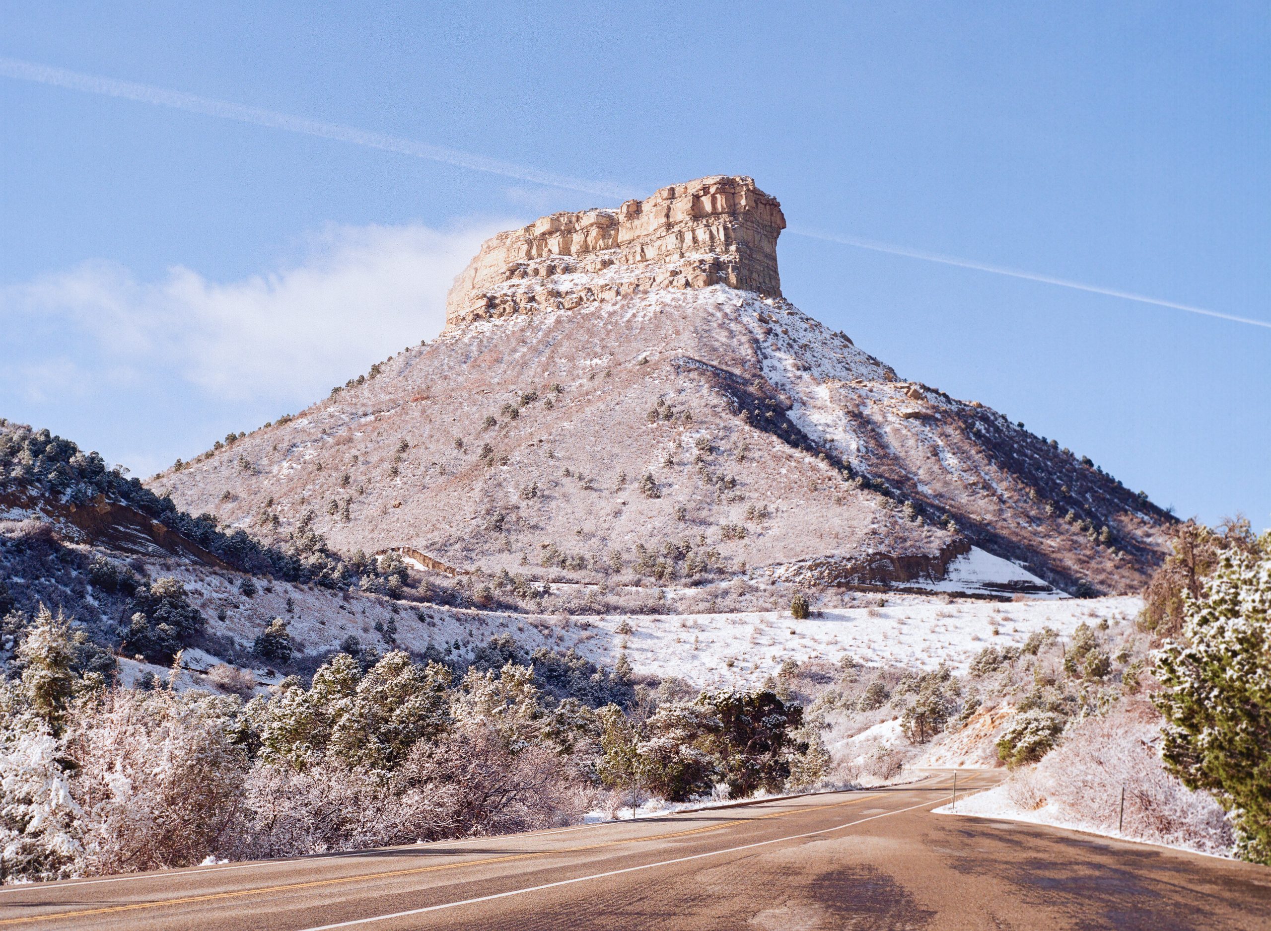 A mesa rises above a snowy landscape, captured in stunning film photography, with a winding road in the foreground and trees scattered along the hillside under a clear blue sky. | Photo by Millworks Film Photography & Prints