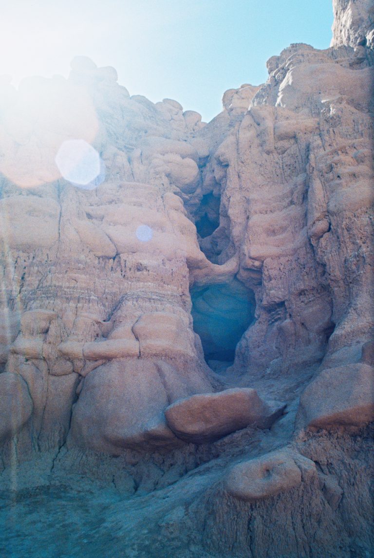 Rocky sandstone formations with eroded shapes and crevices, illuminated by bright sunlight. Captured in the style of film photography, the clear blue sky features lens flare in the upper left corner of this professional scene. | Photo by Millworks Film Photography & Prints