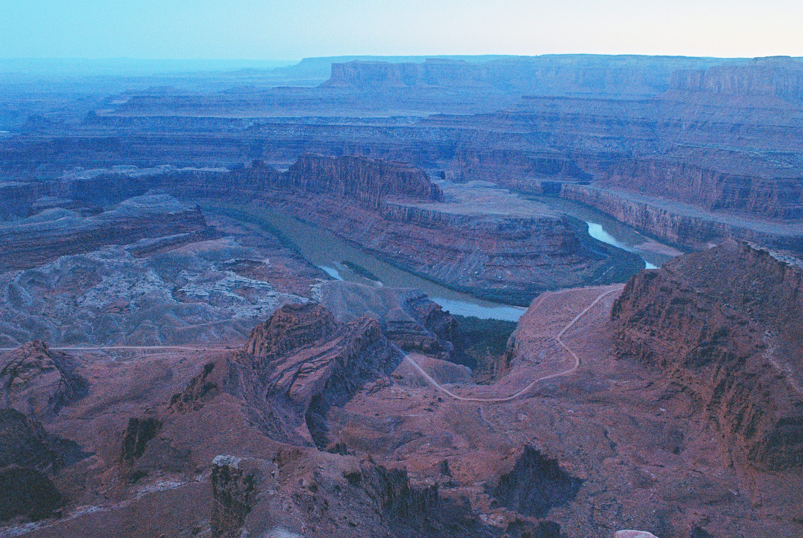 A wide view of a deep canyon with winding river, reddish rocky cliffs, and layered plateaus under soft twilight, captured in stunning professional photography. Dirt roads trace along the canyon’s edge. | Photo by Millworks Film Photography & Prints