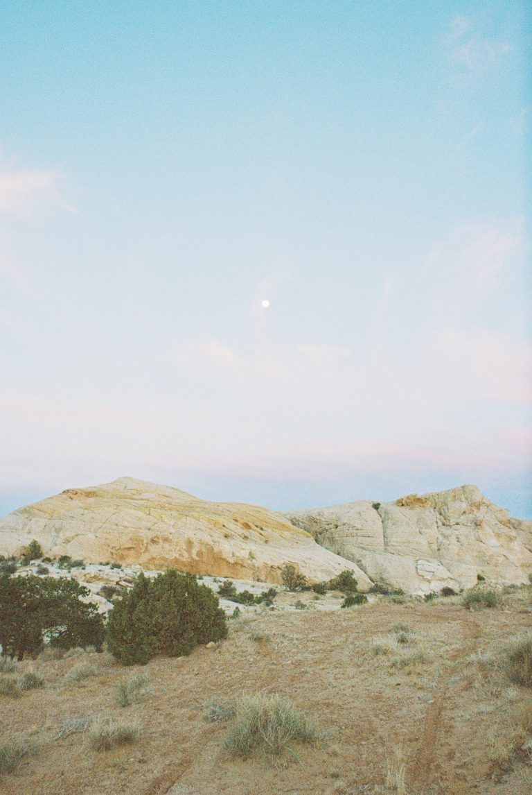 A scenic desert landscape with sandy, rocky hills, scattered shrubs, and a pale blue sky at dusk. The moon is visible above the hills, and soft sunlight gives the rocks a warm, golden tint—captured in the timeless style of film photography. | Photo by Millworks Film Photography & Prints