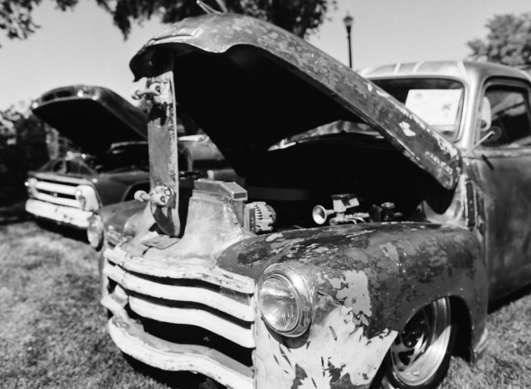 A black-and-white film photo of two vintage cars with weathered exteriors, parked on grass with their hoods open to showcase their engines. Trees and a lamp post are visible in the background. | Photo by Millworks Film Photography & Prints